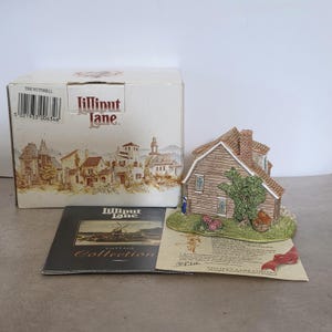 May include: A Lilliput Lane miniature cottage with a brown wooden exterior, a brick chimney, and a green tree. The cottage sits on a green base with flowers. The box and brochure are also visible.