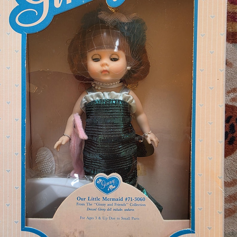 K&ouml;nnte beinhalten: Ginny-Puppe, "Our Little Mermaid #71-3060", in der Originalverpackung. Die Puppe tr&auml;gt ein gr&uuml;nes Paillettenkleid, eine Perlenkette und einen Schleierhut. Sie enth&auml;lt ein rosa Accessoire und stammt aus der "Ginny and Friends"-Kollektion.