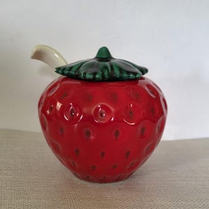 May include: A ceramic strawberry-shaped jar with a green leaf-shaped lid and a small, curved handle. The jar is a vibrant red with small, dark brown speckles, and the lid is a glossy green. The handle is off-white.