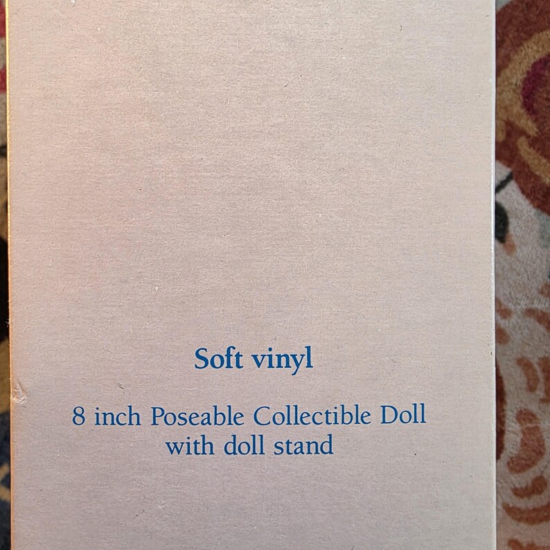 K&ouml;nnte beinhalten: Eine hellblaue Karte mit den Worten "Soft vinyl" und "8 inch Poseable Collectible Doll with doll stand" in blauem Text. Die Karte ist leicht zerknittert.