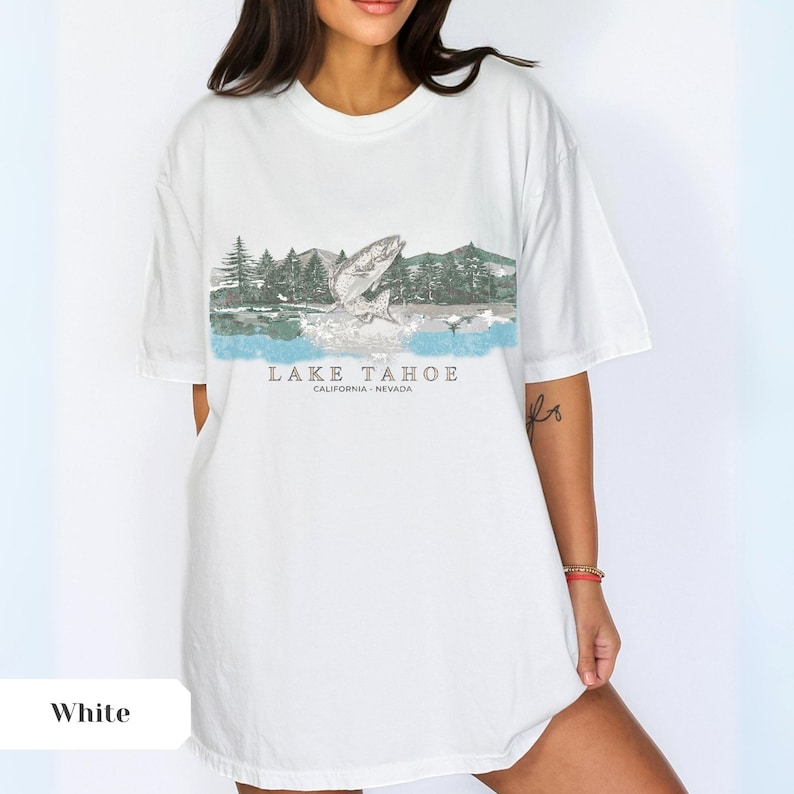 K&ouml;nnte beinhalten: Wei&szlig;es Oversize-T-Shirt mit einem grafischen Aufdruck eines Fisches, der aus dem Wasser springt, mit einer Bergkette und B&auml;umen im Hintergrund. Der Text "LAKE TAHOE CALIFORNIA - NEVADA" ist unter dem Grafikdruck aufgedruckt.