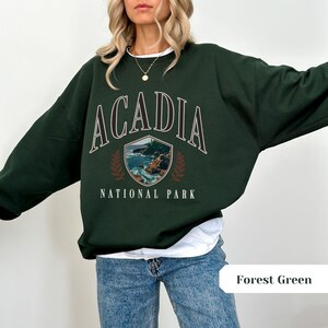 May include: Forest green sweatshirt with the text "ACADIA NATIONAL PARK" in a vintage style. The design includes a shield with a coastal landscape and laurel leaves. The sweatshirt is a relaxed fit.