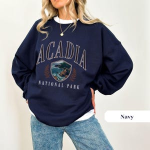 May include: Navy blue sweatshirt with the text "ACADIA NATIONAL PARK" in white and brown. The graphic features a shield with a coastal landscape and laurel leaves. The sweatshirt is a crew neck style.