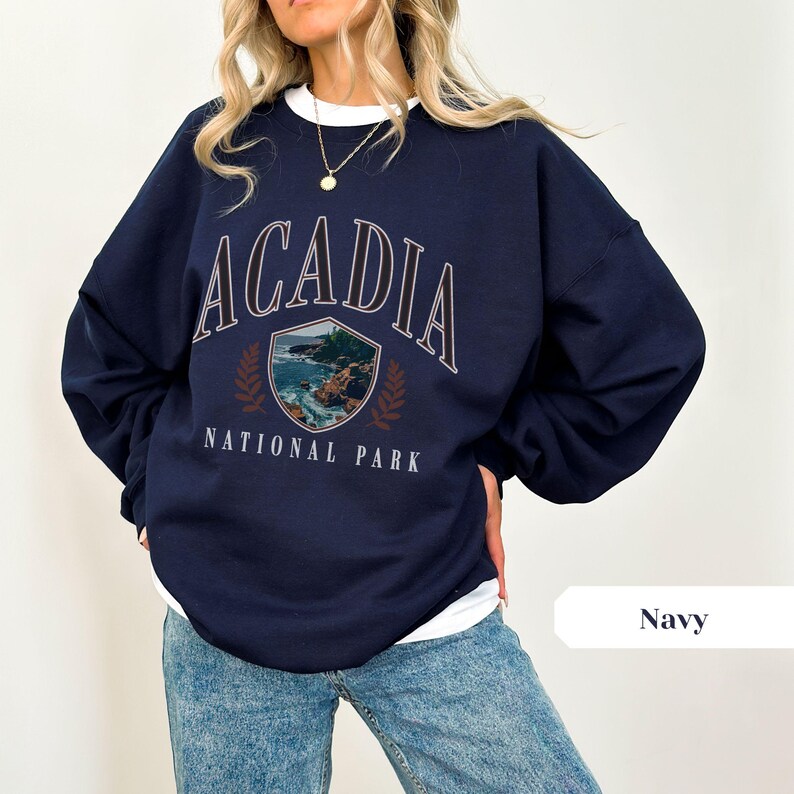 May include: Navy blue sweatshirt with the text "ACADIA NATIONAL PARK" in white and brown. The graphic features a shield with a coastal landscape and laurel leaves. The sweatshirt is a crew neck style.