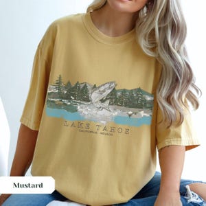 K&ouml;nnte beinhalten: Ein senfgelbes T-Shirt mit einem Aufdruck eines Fisches, der aus dem Wasser springt. Der Aufdruck ist von B&auml;umen und Bergen umgeben. Der Text "LAKE TAHOE CALIFORNIA - NEVADA" ist unter dem Aufdruck gedruckt.