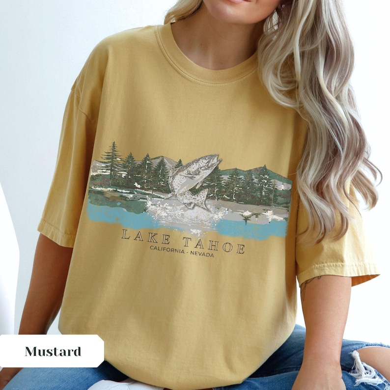 K&ouml;nnte beinhalten: Ein senfgelbes T-Shirt mit einem Aufdruck eines Fisches, der aus dem Wasser springt. Der Aufdruck ist von B&auml;umen und Bergen umgeben. Der Text "LAKE TAHOE CALIFORNIA - NEVADA" ist unter dem Aufdruck gedruckt.