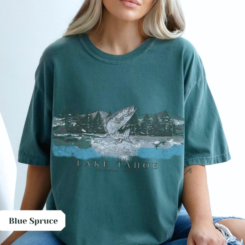 K&ouml;nnte beinhalten: Ein blaugr&uuml;nes T-Shirt mit einem grafischen Aufdruck eines Fisches, der aus dem Wasser springt, vor einer Waldszene. Der Text "LAKE TAHOE CALIFORNIA NEVADA" ist unter dem Grafikdruck aufgedruckt.
