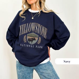 Pu&ograve; includere: Una felpa blu navy con una stampa grafica di un alce davanti a uno scudo con il testo "Yellowstone National Park".