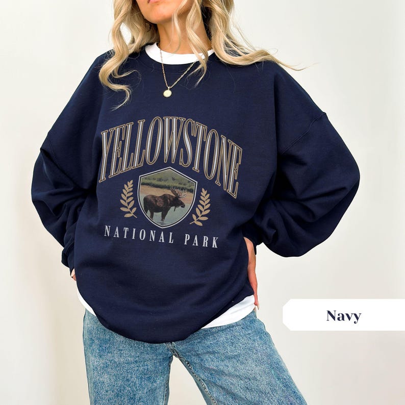 Pu&ograve; includere: Una felpa blu navy con una stampa grafica di un alce davanti a uno scudo con il testo "Yellowstone National Park".