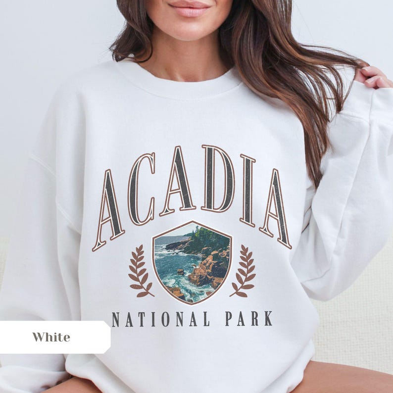 May include: White sweatshirt featuring the text "ACADIA NATIONAL PARK" in a brown, vintage-style font. The design includes a shield with a coastal landscape and laurel leaves. A comfortable, casual top.