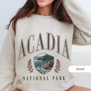 May include: Sand-colored sweatshirt featuring the text "ACADIA NATIONAL PARK" in a vintage style. The design includes a shield-shaped graphic with a coastal landscape and laurel leaves. The sweatshirt is a neutral color.