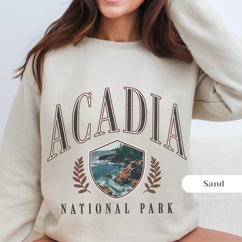 May include: Sand-colored sweatshirt featuring the text "ACADIA NATIONAL PARK" in a vintage style. The design includes a shield-shaped graphic with a coastal landscape and laurel leaves. The sweatshirt is a neutral color.
