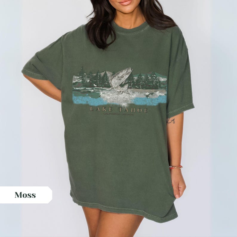 K&ouml;nnte beinhalten: Ein &uuml;berdimensioniertes T-Shirt in Moosgr&uuml;n mit einem grafischen Aufdruck eines Fisches, der aus dem Wasser springt, mit Bergen und B&auml;umen im Hintergrund. Der Text "LAKE TAHOE" ist unter dem Grafikdruck aufgedruckt.