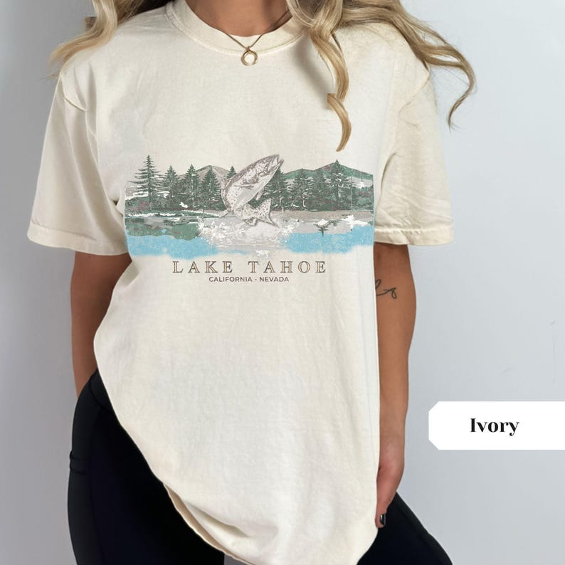 K&ouml;nnte beinhalten: Kurz&auml;rmliges T-Shirt in Elfenbein mit einem grafischen Aufdruck eines Fisches, der aus dem Wasser springt, mit B&auml;umen und Bergen im Hintergrund. Der Text "LAKE TAHOE CALIFORNIA - NEVADA" ist unter dem Grafikdruck aufgedruckt.