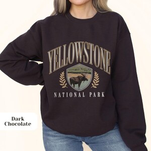Pu&ograve; includere: Una felpa di colore marrone cioccolato scuro con il testo "Yellowstone National Park" in lettere bianche e dorate. Uno scudo con un alce all'interno &egrave; sopra il testo.