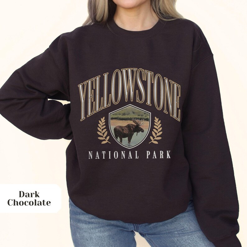 Pu&ograve; includere: Una felpa di colore marrone cioccolato scuro con il testo "Yellowstone National Park" in lettere bianche e dorate. Uno scudo con un alce all'interno &egrave; sopra il testo.