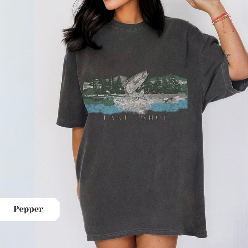 K&ouml;nnte beinhalten: Ein dunkelgraues Oversize-T-Shirt mit einem grafischen Aufdruck eines Fisches, der vor einem Wald aus dem Wasser springt. Der Text "Lake Tahoe" ist unter dem Aufdruck gedruckt.