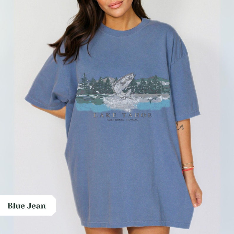 K&ouml;nnte beinhalten: Ein Oversize-T-Shirt aus Jeansblau mit einem grafischen Aufdruck eines Fisches, der aus dem Wasser springt, mit einem Berg- und Waldhintergrund. Der Text "LAKE TAHOE CALIFORNIA - NEVADA" ist unter dem Grafikdruck aufgedruckt.