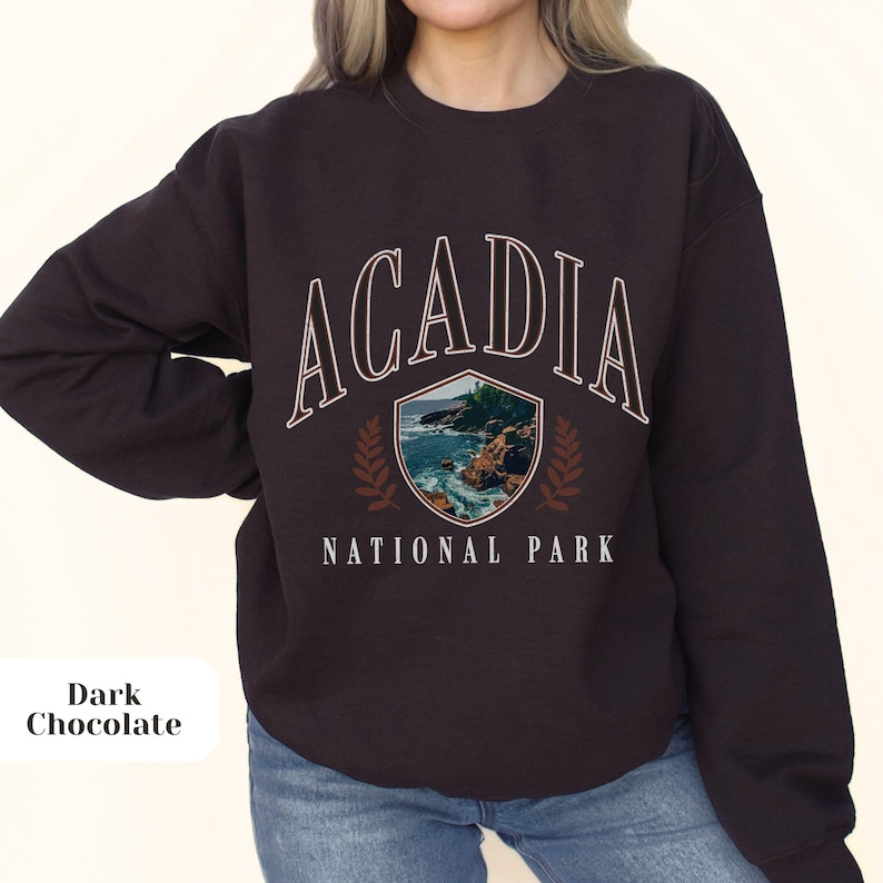 May include: Dark chocolate-colored sweatshirt with the text "ACADIA NATIONAL PARK" in white. The design includes a shield with a coastal scene and laurel branches. The sweatshirt is a comfortable, casual style.