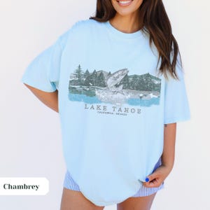 K&ouml;nnte beinhalten: Ein hellblaues T-Shirt mit einem grafischen Aufdruck eines Fisches, der aus dem Wasser springt, mit Bergen und B&auml;umen im Hintergrund. Der Text "LAKE TAHOE CALIFORNIA - NEVADA" ist unter dem Grafikdruck aufgedruckt.