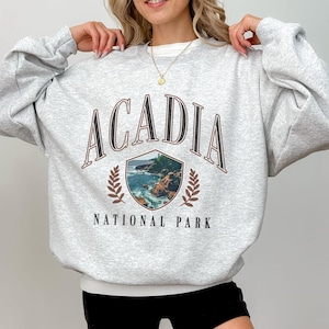 May include: A light gray sweatshirt with the word "ACADIA" arched across the top in a brown font. Below the word is a shield-shaped graphic of a coastal scene. The words "NATIONAL PARK" are printed below the graphic.