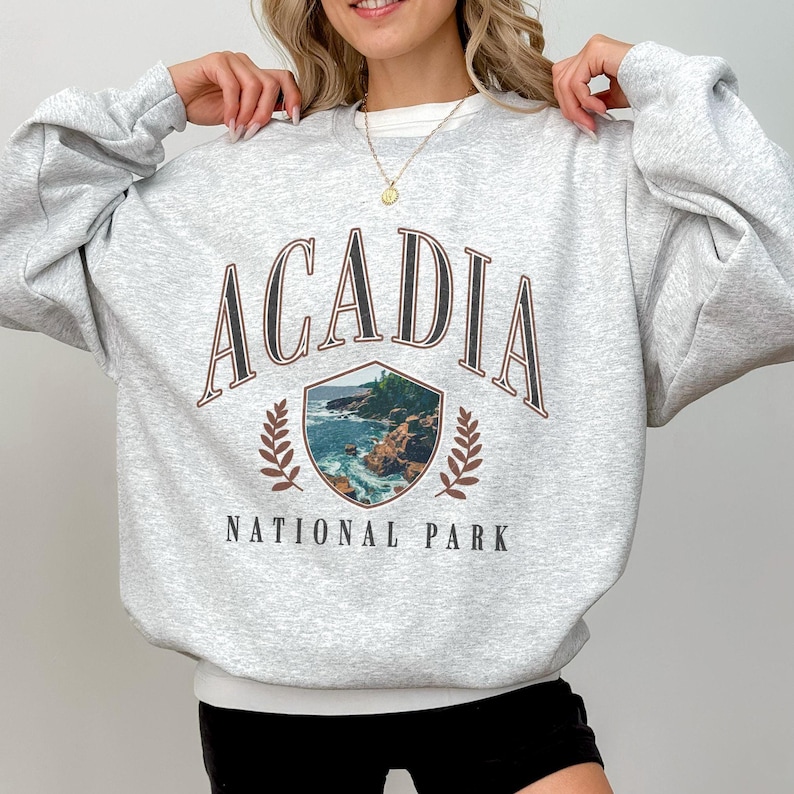 May include: A light gray sweatshirt with the word "ACADIA" arched across the top in a brown font. Below the word is a shield-shaped graphic of a coastal scene. The words "NATIONAL PARK" are printed below the graphic.