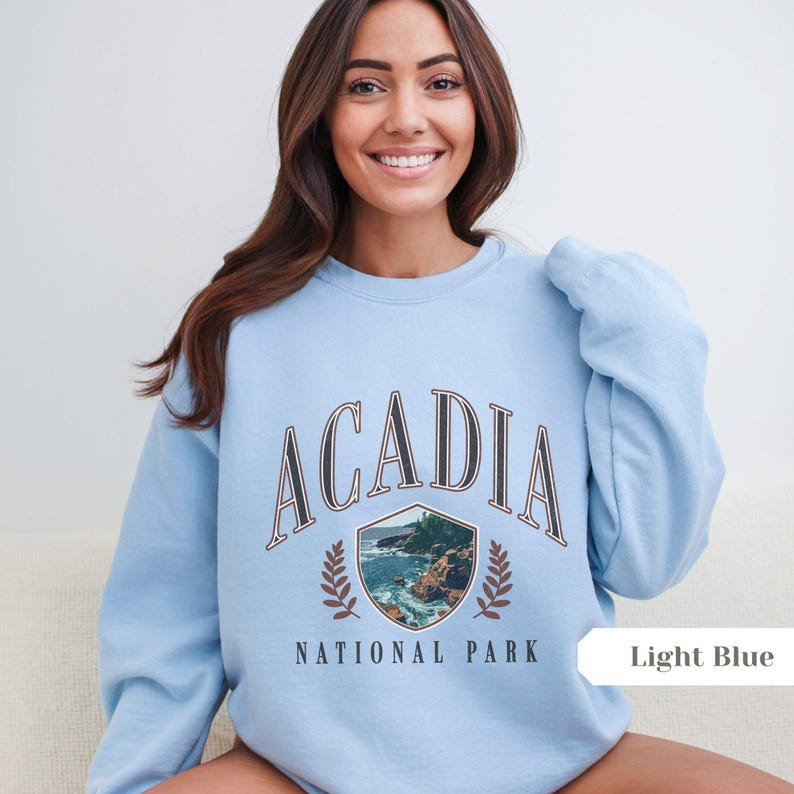 May include: Light blue sweatshirt with the word "ACADIA" in a vintage font. Below the word is a shield-shaped graphic with a coastal scene and the words "NATIONAL PARK". The sweatshirt is a light blue color.