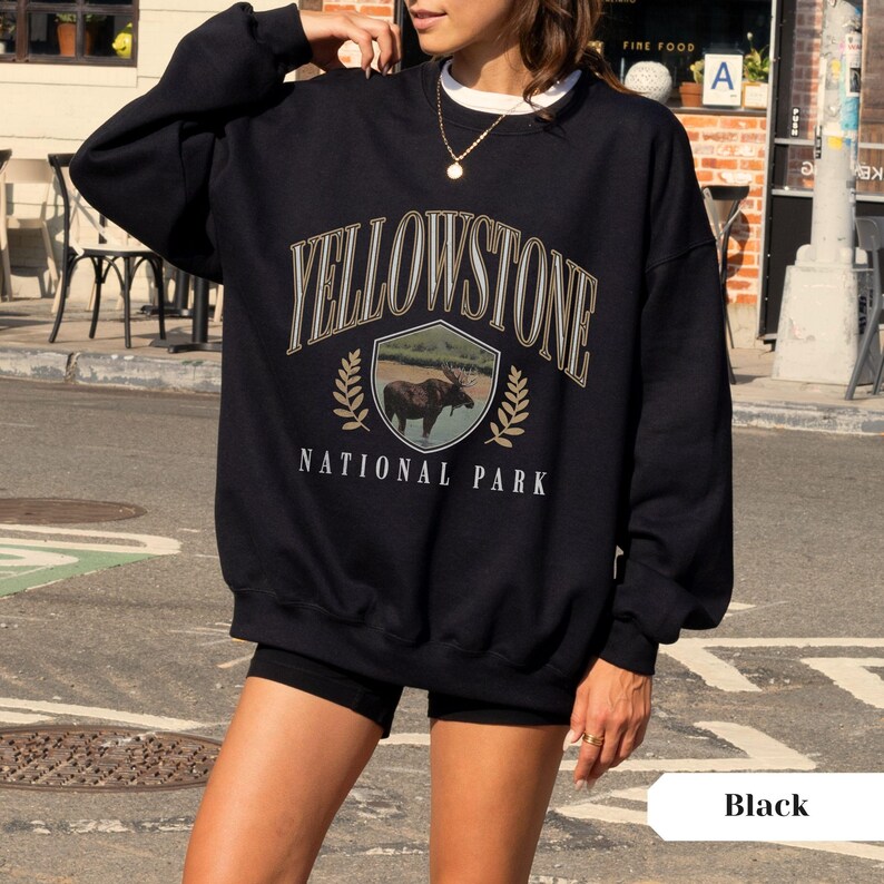 Pu&ograve; includere: Felpa nera con una stampa grafica di un alce in uno scudo con il testo "Yellowstone National Park" in un font di stile vintage.