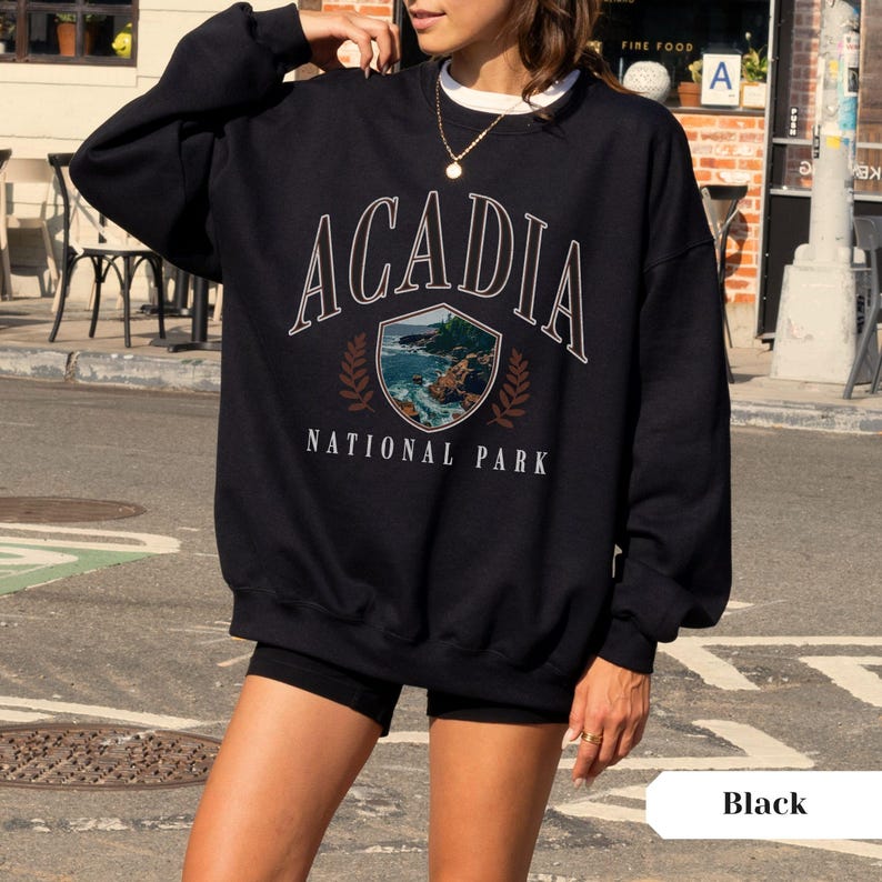 May include: Black sweatshirt with the text "ACADIA NATIONAL PARK" in white lettering. The design includes a shield-shaped graphic with a coastal landscape and brown leaf accents. The sweatshirt is a crew neck style.