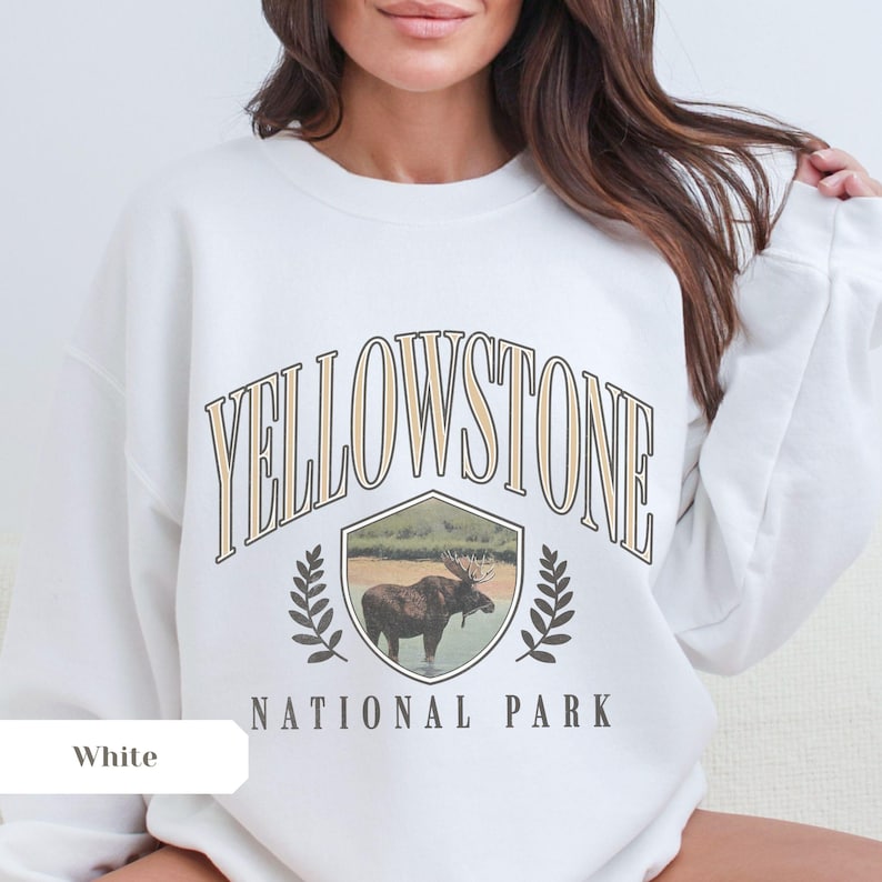 Pu&ograve; includere: Felpa bianca con una stampa grafica di un alce davanti a una scena di foresta. Il testo "Yellowstone National Park" &egrave; stampato in un font di stile vintage.