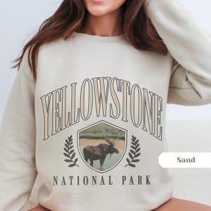 Pu&ograve; includere: Una felpa beige con il testo "YELLOWSTONE NATIONAL PARK" in un font di stile vintage. La grafica presenta un alce in uno scudo con uno sfondo di foresta. La parola "Sand" &egrave; scritta in un riquadro bianco nell'angolo in basso a destra.