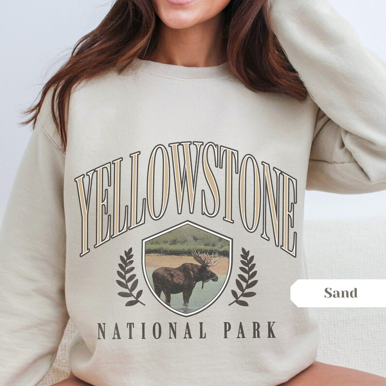 Pu&ograve; includere: Una felpa beige con il testo "YELLOWSTONE NATIONAL PARK" in un font di stile vintage. La grafica presenta un alce in uno scudo con uno sfondo di foresta. La parola "Sand" &egrave; scritta in un riquadro bianco nell'angolo in basso a destra.