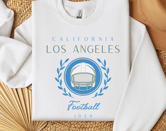 May include: White sweatshirt with a blue and white football helmet graphic inside a blue circle and laurel wreath. The text "CALIFORNIA LOS ANGELES Football 1959" is printed above and below the graphic. The sweatshirt is made of a soft material.