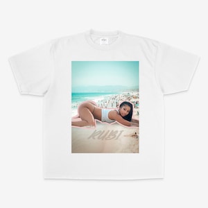 May include: White t-shirt featuring a beach scene with a person lying on the sand. The word "RUBI" is printed on the sand. The beach scene includes a blue ocean, sky, and a crowd of people.
