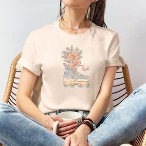 May include: Cream-colored t-shirt featuring a pastel illustration of a roller skate filled with flowers and a bow. The roller skate is light blue with peach and yellow accents. The flowers are peach and the bow is peach with flowing ribbons.