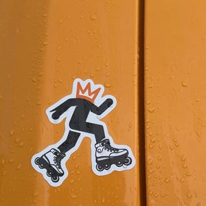 May include: A sticker featuring a black silhouette of a person wearing roller skates and a crown. The sticker is on an orange surface with water droplets.