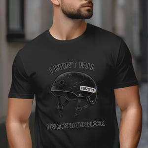 May include: Black t-shirt with the text "I DIDN'T FALL I BLOCKED THE FLOOR" above and below a distressed black helmet graphic. The helmet has a white label that says "TEEZLYPR".