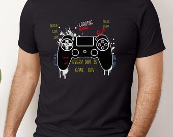 May include: Black t-shirt featuring a white graphic of a video game controller with the text "EVERY DAY IS GAME DAY." Additional text includes "NEVER GIVE UP!" and "PRESS START." The design has a splatter effect.