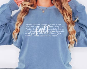 May include: A light blue sweatshirt with the word "fall" in white text. The sweatshirt has a list of fall-themed words and phrases printed on it, including "golden leaves", "cinnamon", "warm hugs", "pumpkin spice", "apple cider", "crisp air", "fluffy scarves", "cozy sweaters", "bonfire nights", "football", "give thanks", "corn maze", "turkey", "pie", "crispy leaves", "candy corn", and "jeans".
