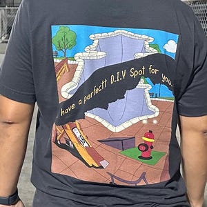 Può includere: Una maglietta nera con una stampa grafica di uno skatepark. La grafica presenta una half-pipe, una rampa e un idrante. Il testo "I have a perfect D.I.Y Spot for you." è stampato sulla grafica.
