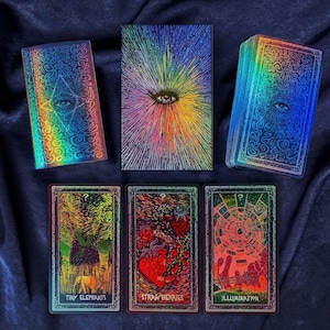Op de afbeelding: Een set tarotkaarten met holografische en kleurrijke ontwerpen. De set bevat kaarten met afbeeldingen van een oog, kleine olifanten, aardbeien en een verlicht ontwerp. De kaarten zijn gerangschikt op een donkerblauwe stof.