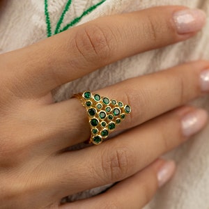 Puede incluir: Un anillo de oro con un diseño triangular que presenta un grupo de piedras preciosas verdes.