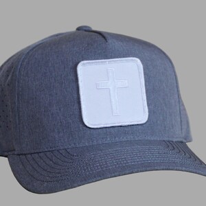 May include: A gray baseball cap with a white square patch featuring a cross design. The cap has a curved brim and small ventilation holes on the side. The hat is made of a textured fabric.