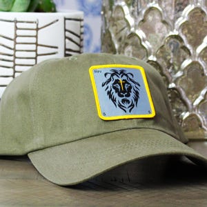 May include: Olive green baseball cap with a lion patch. The patch has a lion's head design with the word "Rev" at the top. The cap is made of a soft material and has a curved brim.