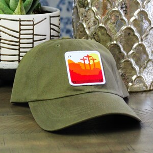 May include: A green baseball cap with a patch featuring a red, orange, and yellow sunset with three crosses. The patch has the text "LK 23:33-43" on it.