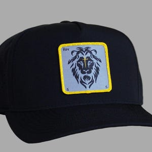 May include: Black baseball cap with a rectangular patch featuring a lion's head design. The patch has a light blue background with a yellow border and the text "Rev" above the lion. The number "5" is at the bottom corners of the patch.