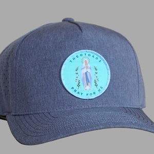 May include: A blue baseball cap with a curved brim. The cap features a circular patch with the text "THEOTOKOS PRAY FOR US" surrounding an illustration of a religious figure. The cap has small ventilation holes.