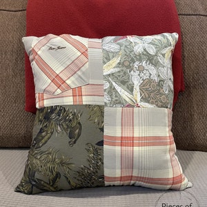 May include: Square decorative pillow with a patchwork design. The pillow features four distinct fabric patterns: plaid, floral, and tropical leaf prints in shades of gray, red, and green. The pillow is resting on a brown sofa with a red pillow behind it.