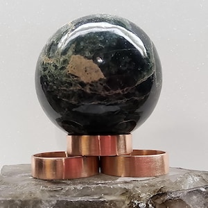 May include: A dark green and black sphere sits atop three copper-colored rings. The sphere has a marbled appearance with lighter brown and green patches. The rings are evenly spaced and have a matte finish. The objects rest on a rough, gray surface.