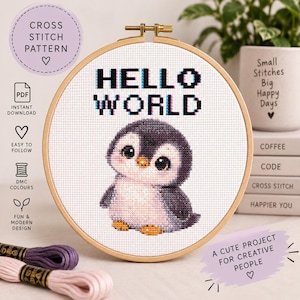 Op de afbeelding: Een kruissteekpatroon in een houten borduurring met de tekst HELLO WORLD in een zwart pixellettertype boven een kleine, ronde, grijs-witte pinguïn met grote ogen en oranje voeten op een witte stoffen achtergrond.